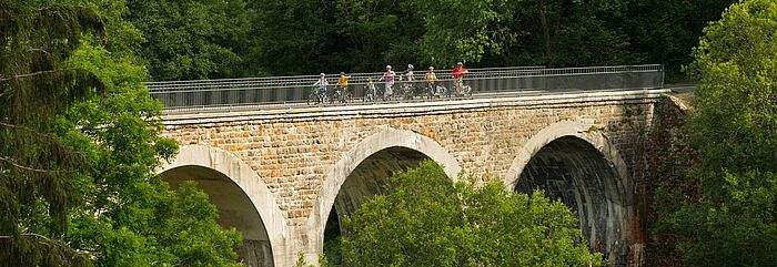 Man sieht eine alte große Brücke auf der eine Familie Fahrrad fährt.