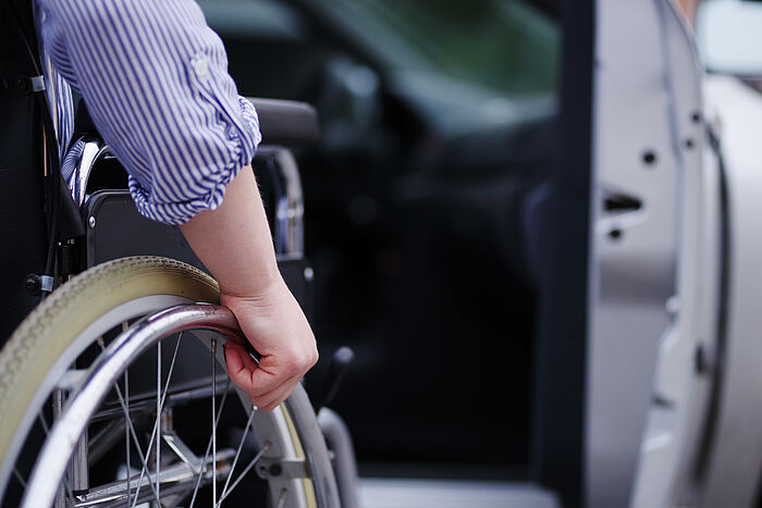 Jemand in einem Rollstuhl bewegt sich auf die offene Türe eines silbernen Fahrzeugs zu. Zu sehen ist von der Person nur der rechte Arm und die obere Hälfte des rechten Rades.