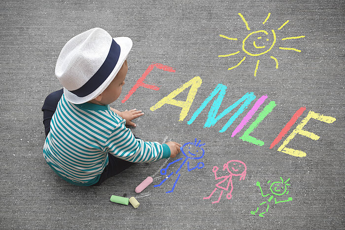 Ein Kind mit Hut malt mit bunten Kreidestifte das Wort Familie und Strichmännchen auf den Asphalt