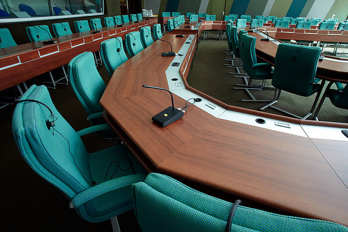 Ausschnitt eines Sitzungssaales, grüne/türkise Stühle kreisförmig in mehreren Reihen an braunen Tischen mit Mikrofonen und Headsets