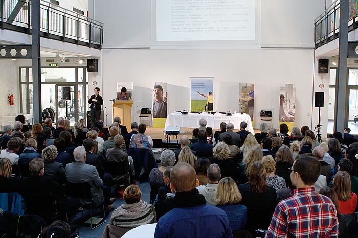 Publikum und Podium mit Vortragenden und einer Gebärdendolmetscherin