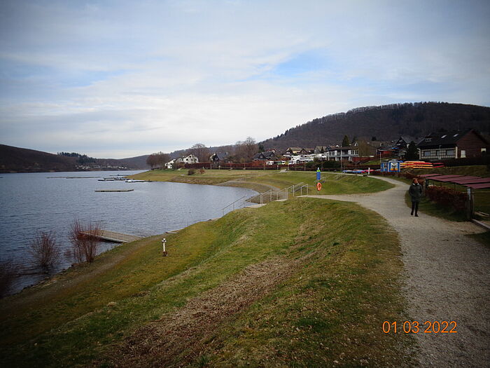 Eine Bucht mit Wasser auf der linken Seite, Weg und Grünflächen auf der rechten Seite und im Hintergrund Hügel, der Himmel ist blau mit Wolken