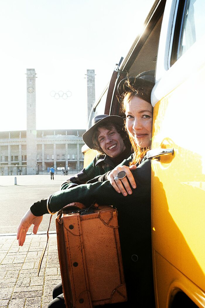 Die Sängerin Magdalena Ganter und Max Prosa sitzen in der Seitentür eines gelben VW Bullis, einen Koffer auf den Knien. Im Hintergrund ist das Berliner Olympiastadion zu sehen.
