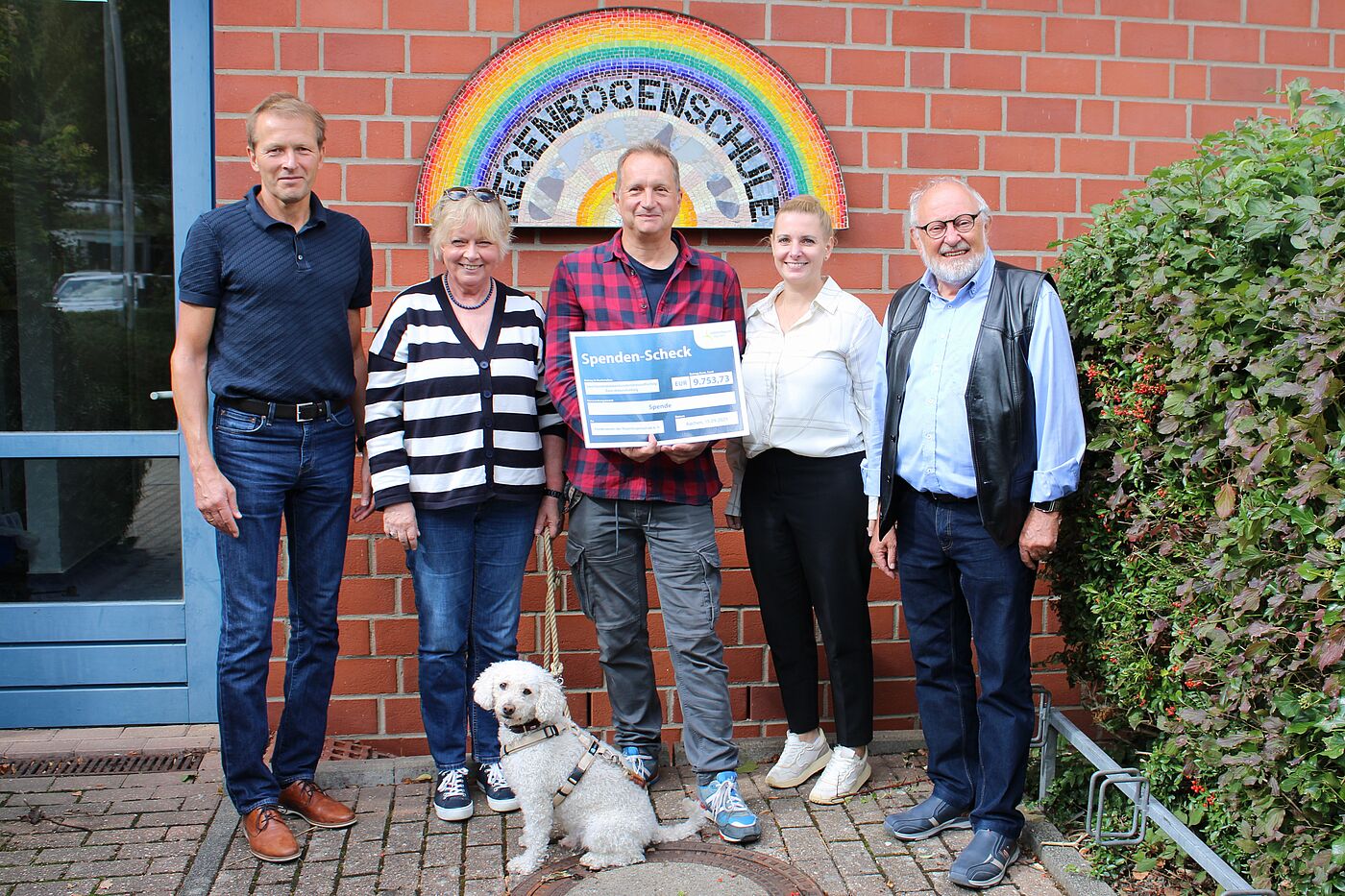 Drei Männer, zwei Frauen und ein Hund vor einem Regenbogen-Schild mit Spendenscheck in der Hand.