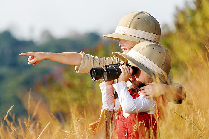 Kinder mit Safari-Hüten schauen durch ein Fernglas und zeigen auf "Aktuelles"
