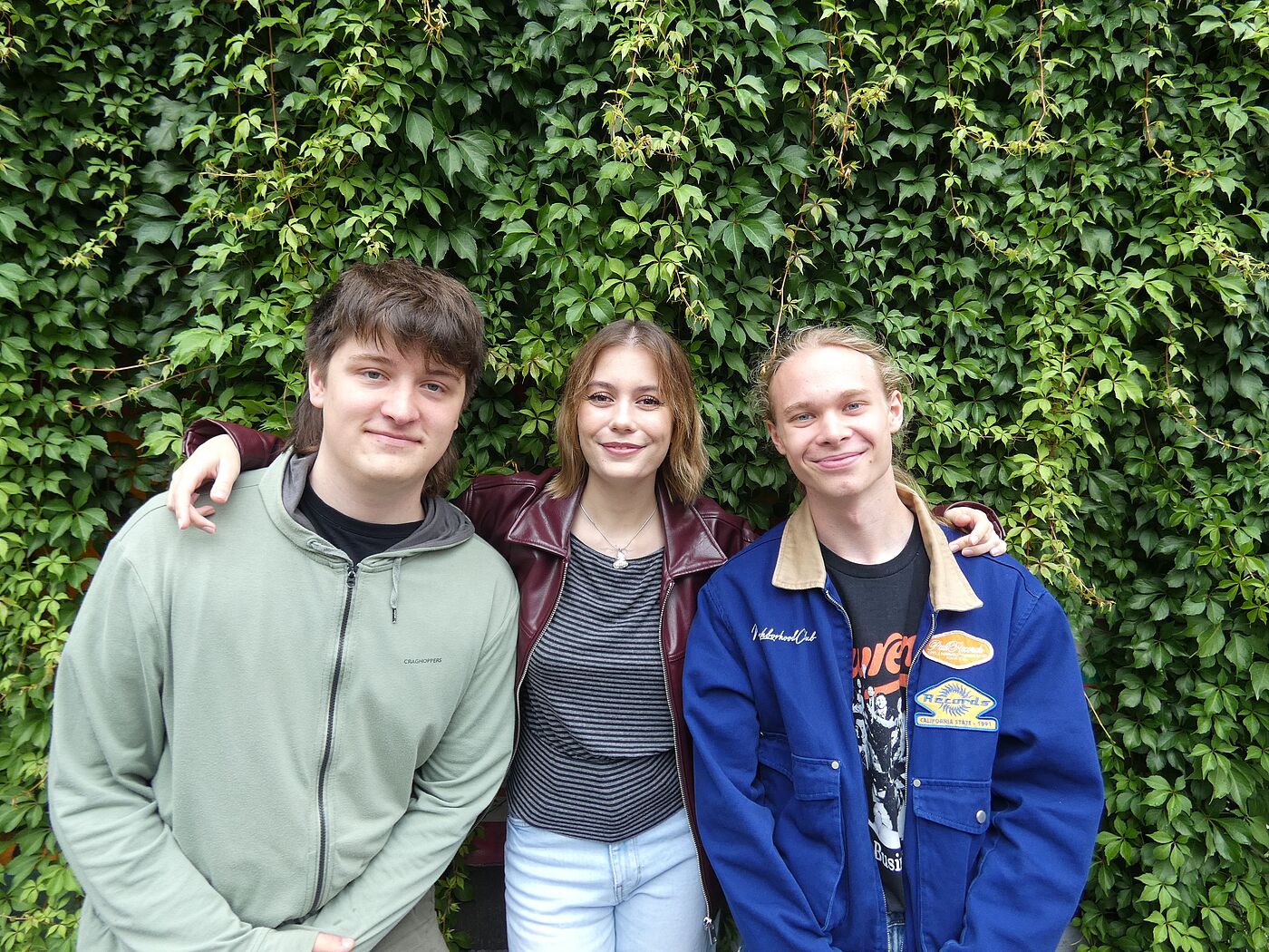 Ein junger Mann und zwei junge Frauen vor einer Hecke.