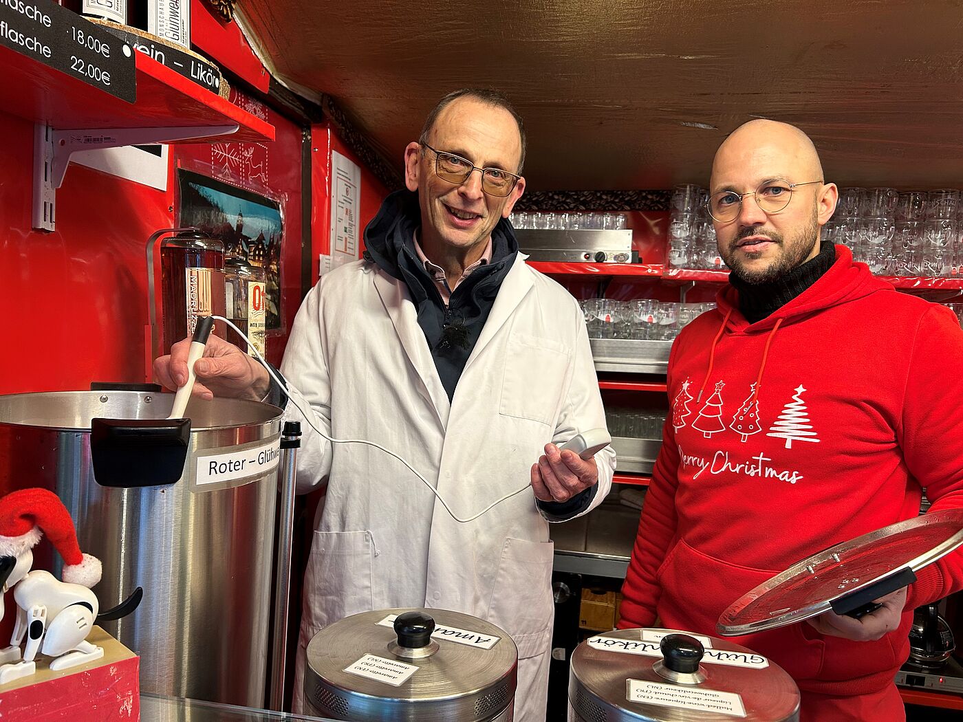 Ein Mann mit Kittel und ein Mann in Weihnachtspullover. Der Mann im Kittel hält ein Thermometer in einen Glühweinbehälter.