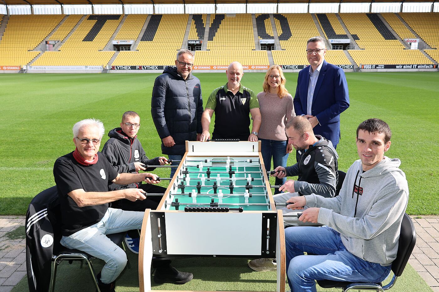 Vier Männer sitzen an einem Tischkicker. Dahinter stehen drei Männer und eine Frau. Ganz hinten ist eine Fußballtribühne zu sehen.