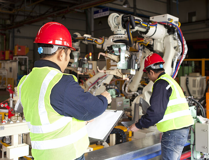 zwei Männer mit gelben Warnwesten und roten Helmen vor einer Industriemaschine. Der eine Mann hält eine Klemmbrett in der Hand und notiert etwas, der andere Mann überprüft die Maschine.