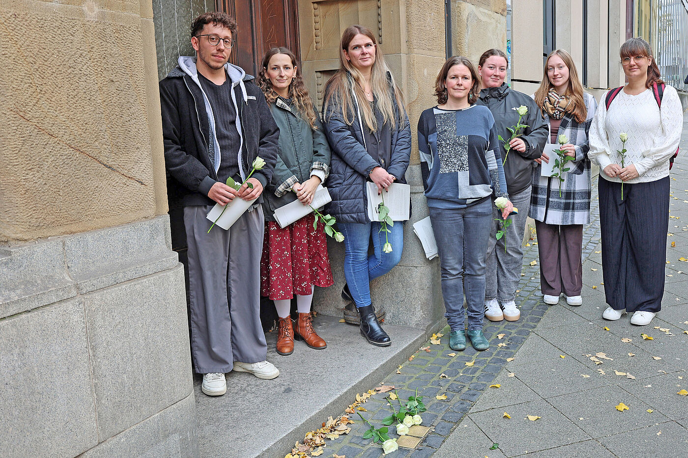 Mehrere junge Menschen vor Stolpersteinen mit weißen Rosen.