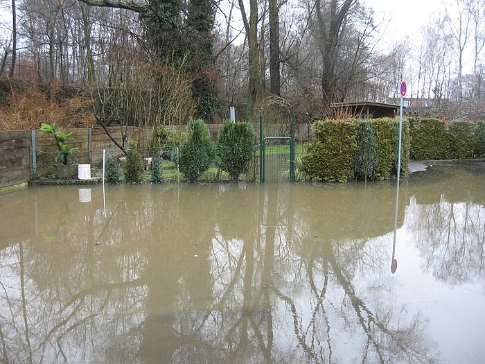 Hochwasser, eine Straße vor einem Garten ist überschwemmt mit Wasser. Das Wasser reicht in die Gartenanlage rein. Im Hintergrund sind Hecken, Bäume, ein Zaun und ein Gartenhäuschen zu sehen.