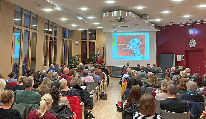 Mehrere Menschen sitzen in einem Saal vor einer Leinwand