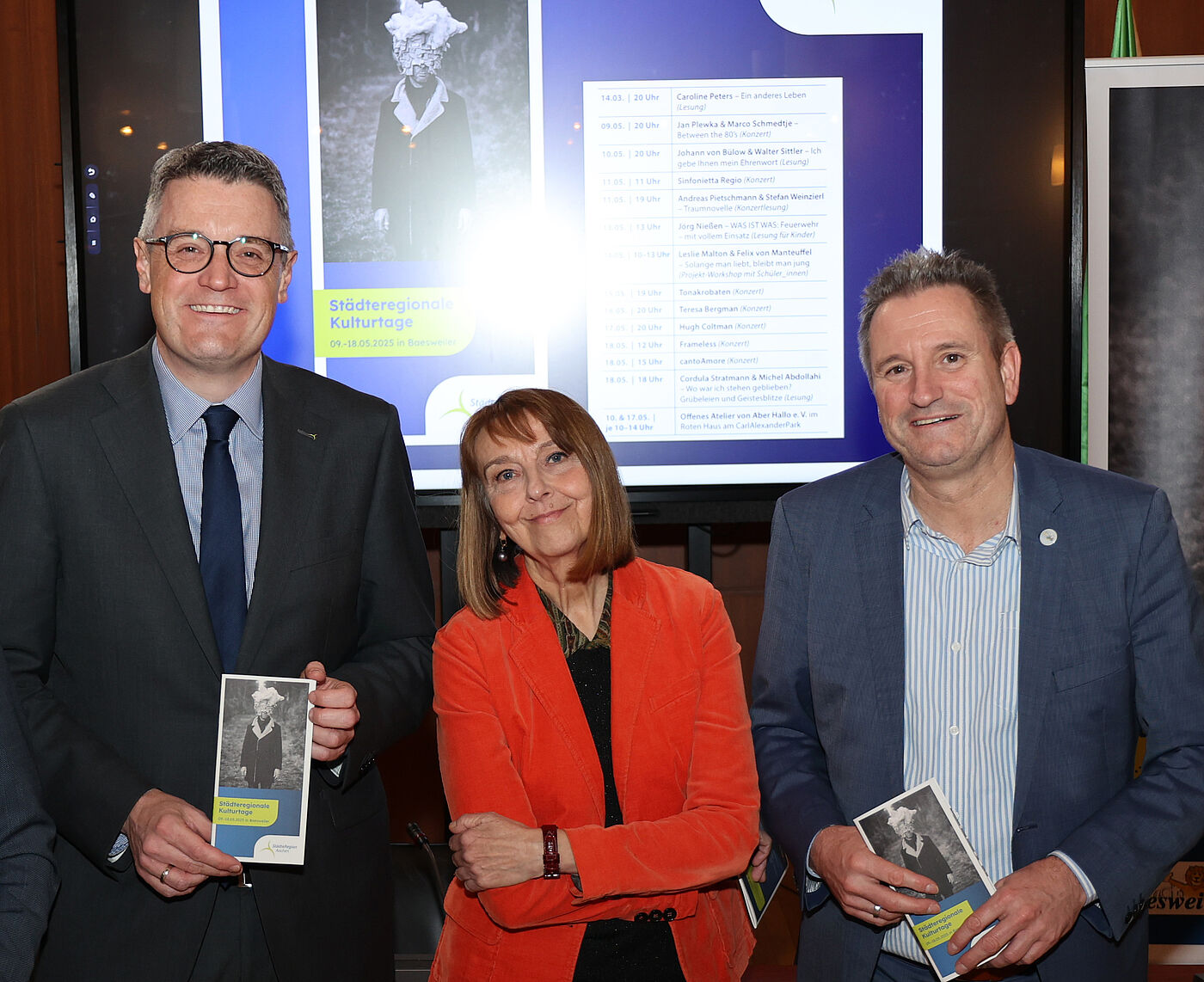 Dr. Tim Grüttemeier, Dr. Nina Mika-Helfmeier sowie Pierre Froesch stehen vor einer Bildschirmpräsentation und halten das Programmheft der Städteregionalen Kulturtage 2025 in den Händen