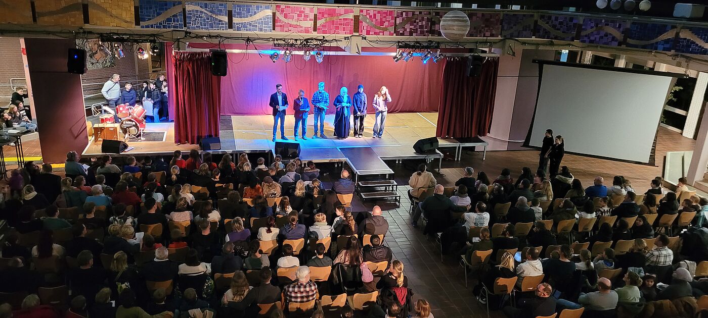 Ein großer Raum voller Menschen mit einer Bühne auf der junge Menschen stehen.