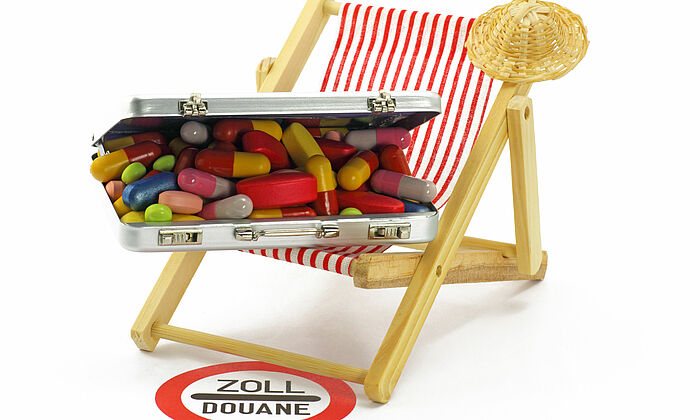 Liegestuhl mit aufgeklapptem Koffer voller Tabletten davor ein Warnschild mit Aufschrift „Zoll/Douane“.