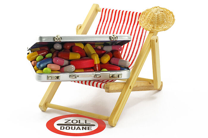 Liegestuhl mit aufgeklapptem Koffer voller Tabletten davor ein Warnschild mit Aufschrift „Zoll/Douane“.