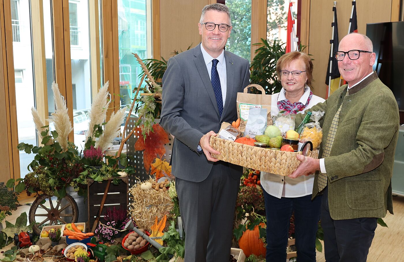 Zwei Männer und eine Frau mit einem herbstlichen Präsentkorb in der Hand vor herbstlich geschmücktem Hintergrund.