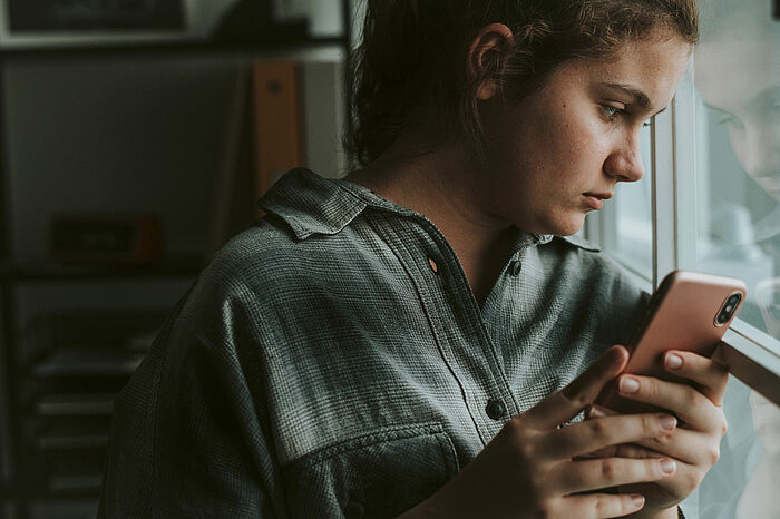 Ein Mädchen mit einem Smartphone in der Hand schaut traurig aus dem Fenster