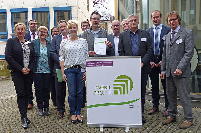 Gruppenfoto. Mehrere Personen stehen hinter einem Schild mit dem Logo von Mobil.Pro.Fit. Eine männliche Person hält eine Mobil.Pro.Fit Auszeichnung in der Hand.