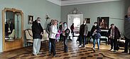 Schloss Wernerdorf: ein Raum mit Parkettboden, Spiegel und Bildern an der Wand, den einige Besuchende besichtigen 