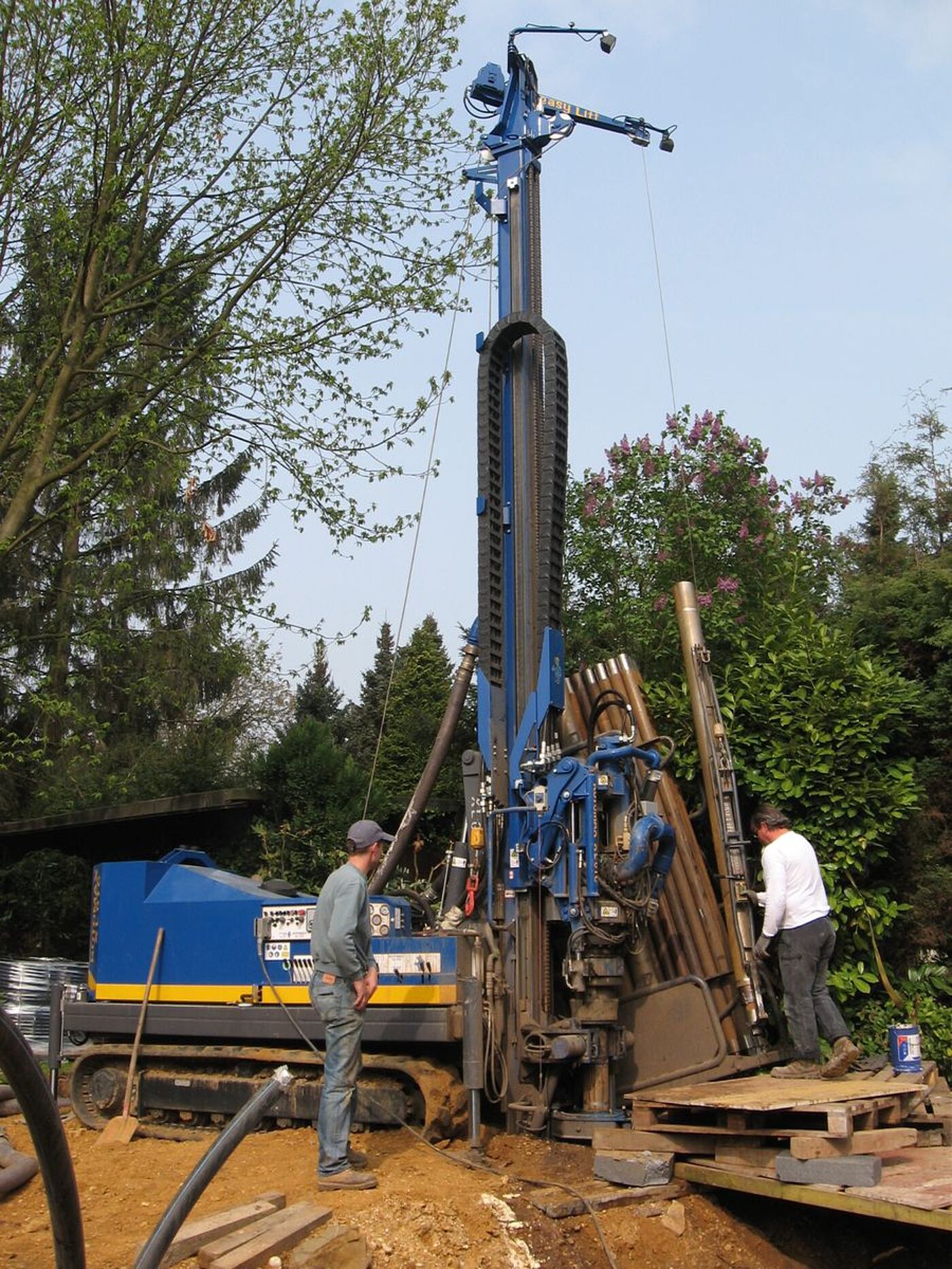 Zu sehen ist, wie zwei Männer mit einem Bohrgerät ein tiefes Loch bohren. Gerade wird ein Bohrstange mit einem Seilzug hochgezogen. 