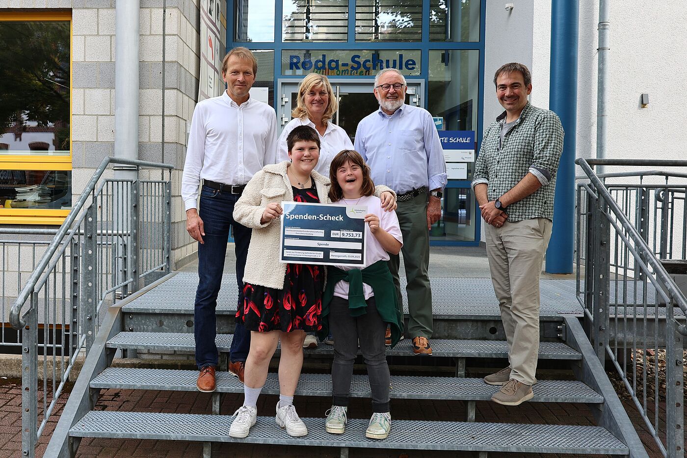 Vier Erwachsene und zwei Jugendliche auf der Treppe vor einer Schule. Sie halten einen Spendenscheck.