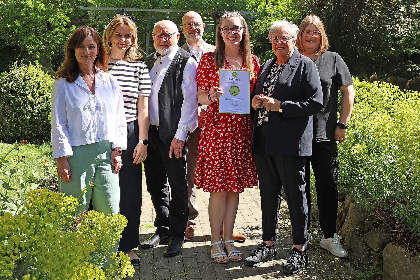 Mehrere Menschen in einem Garten. Eine Frau hält ein Blatt Papier fest.