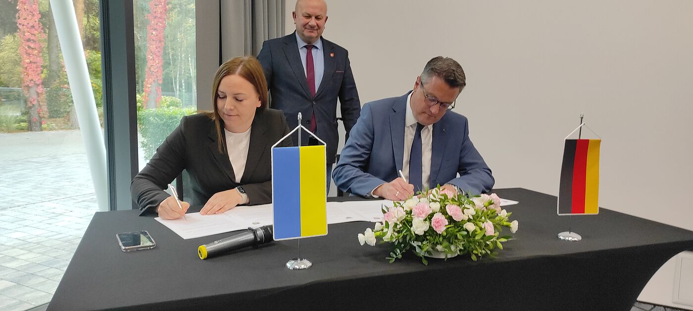 Die Landrätin des Rajon Lwiw, Khrystyna Zamula, und StädteRegionsrat Dr. Tim Grüttemeier sitzen an einem Tisch und unterzeichnen das Partnerschaftsabkommen. Im Hintergrund steht der Landrat des Kreises Lubaczów, Zenon Swatek.