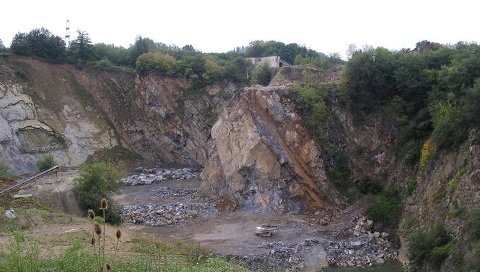 Ein Steinbruch von oben fotographiert. Zu sehen ist eine Brechermaschine und ein See und dicke Felsbrocken. 