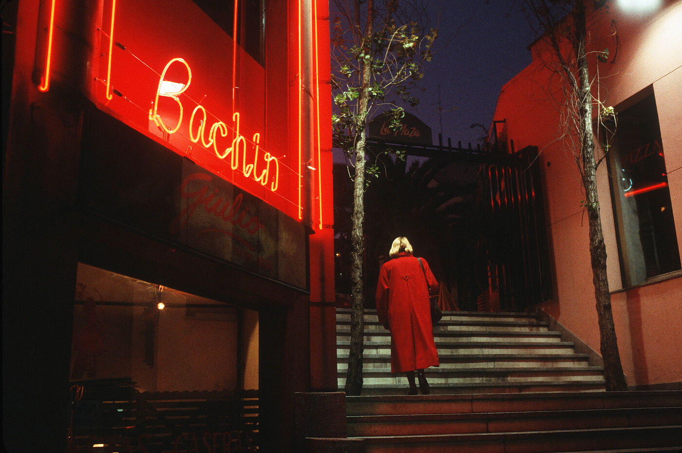 Eine Frau mit rotem Mantel läuft eine Treppe hinauf. Der Hintergrund ist dunkel, sodass die rote Leuchtschrift an einer Hausfassade besonders leuchtet. 