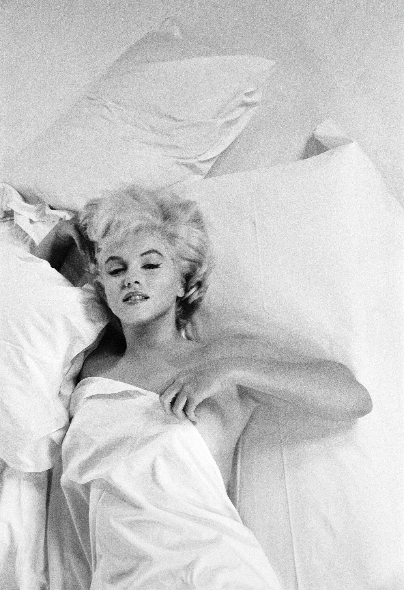 Die Schauspielerin Marilyn Monroe räkelt sich auf einem Bett. Ihr Körper ist durch eine weiße Bettdecke verdeckt, ihr Haar liegt aufgefächert auf der Matratze.