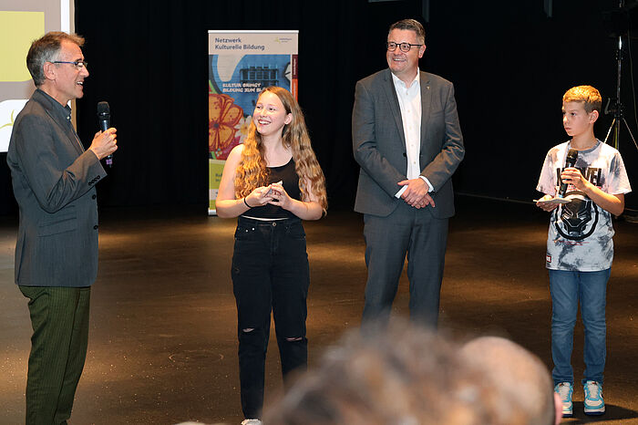 Die jungen Moderatoren Fiona und Tobias führen auf einer Bühne ein Gespräch mit Heinrich Brötz und Tim Grüttemeier