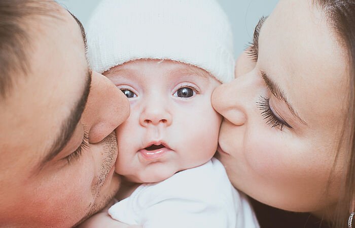 Ein Baby wird von seiner Mutter und seinem Vater auf die Wange geküsst