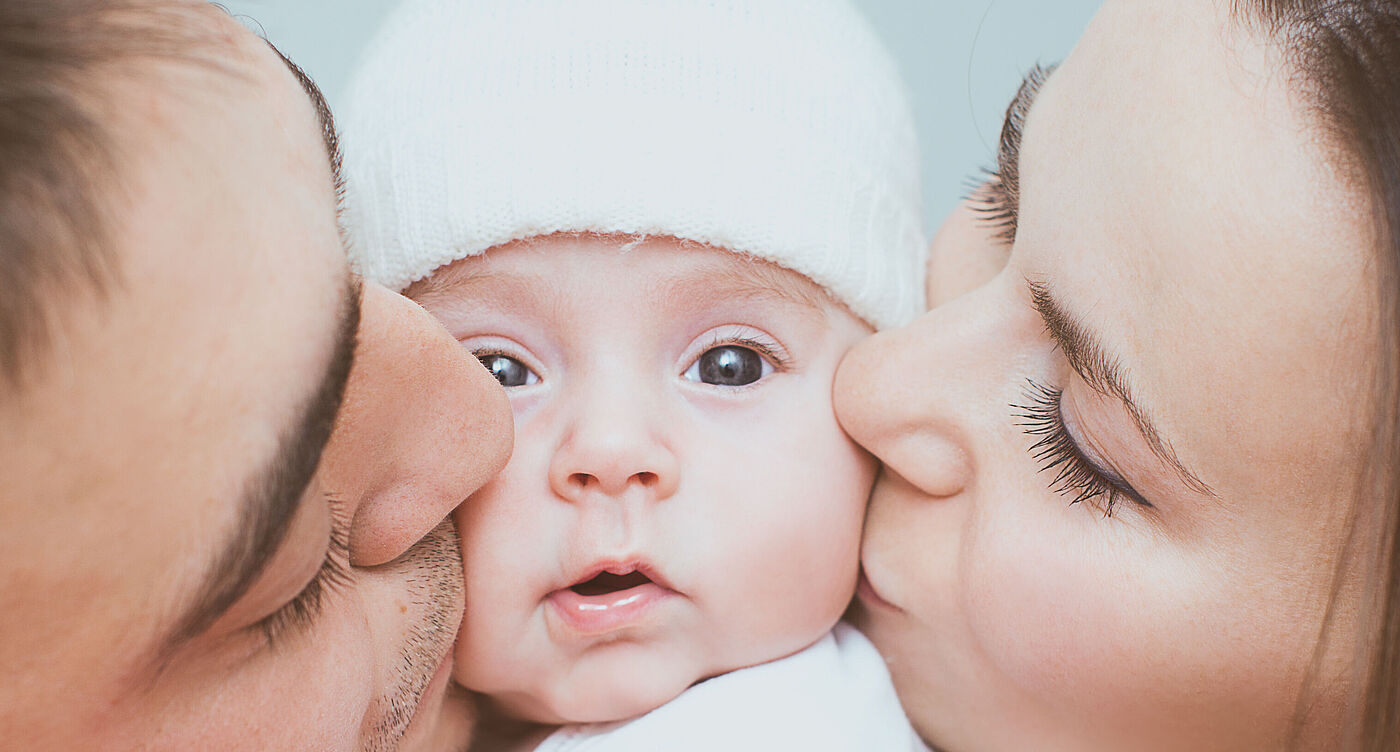 Ein Baby wird von seiner Mutter und seinem Vater auf die Wange geküsst