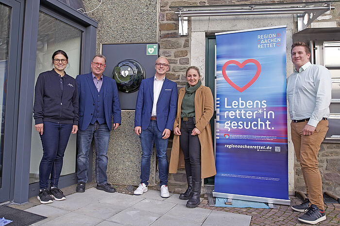 Die INhaber der Firma AixTEC Components zeigen den Defibrillator, den sie so montiert haben, dass er öffentlich erreichbar ist. Mit auf dem Bild sind außerdem Iris Bornewasser (Amt für Rettungswesen und Bevölkerungsschutz der StädteRegion Aachen), Thomas Rissmayer (Inhaber AixTEC Components GmbH), Leon Rissmayer, Ines Krasny (Ärztliche Leitung Rettungsdienst StädteRegion Aachen und Leitung Notarztstandort Stolberg) und Simon Schröder (Leiter Rettungsdienst und Leitstelle im Amt für Rettungswesen und Bevölkerungsschutz der StädteRegion Aachen).