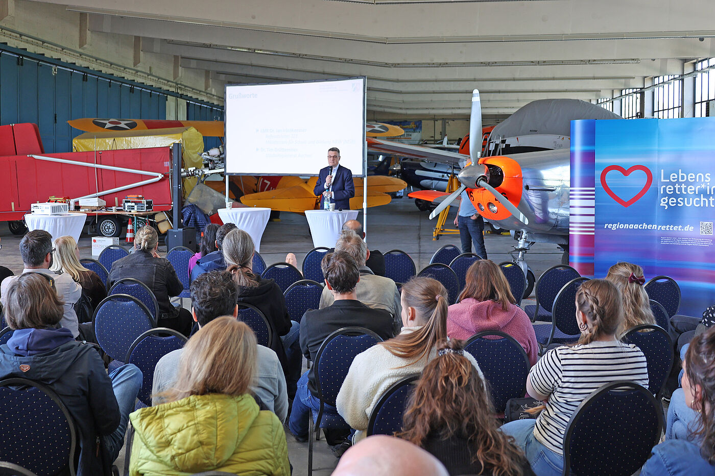 Viele Menschen sitzen in einem Flugzeughangar. Vorne spricht ein Mann zu ihnen.