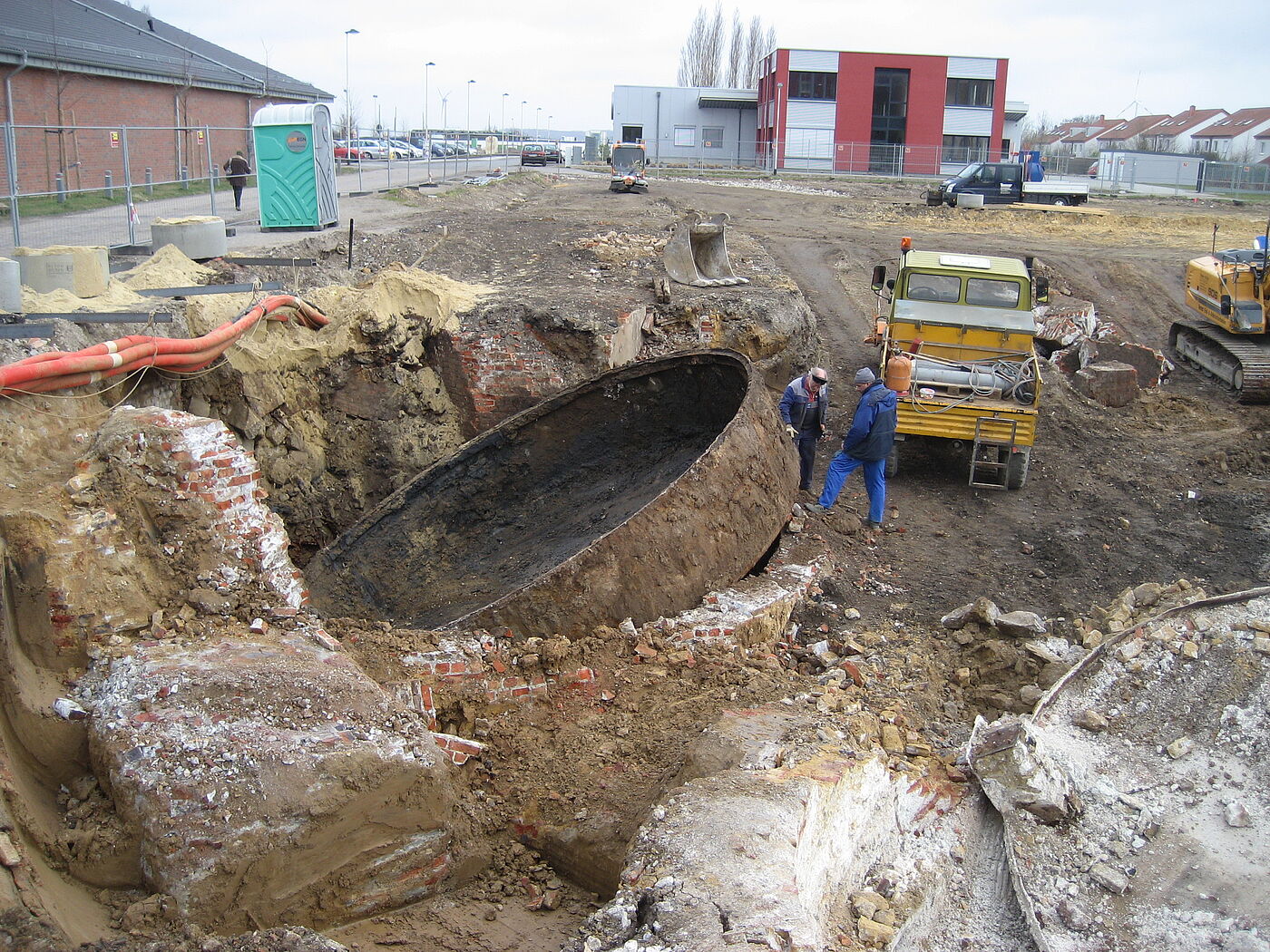 Auf einer Baustelle wird gerade ein riesiges rundes Becken ausgehoben. Zu sehen sind zwei Männer in blauer Kleidung, Baufahrzeuge und im Hintergrund Häuser, Gebäude, parkende Autos vor einem Supermarktgebäude.