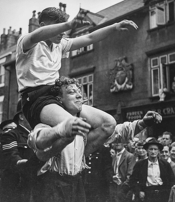 Wolf Suschitzky, Miner's Gala, Durham, 1952 Zwei junge Männer befinden sich während einer Demonstration in den Straßen von Durham. Einer sitzt bei dem anderen auf die Schultern und reisst die Arme nach oben.