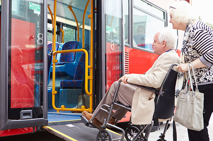 Ältere Frau schiebt älteren Mann im Rollstuhl über eine Rampe in den Bus. 