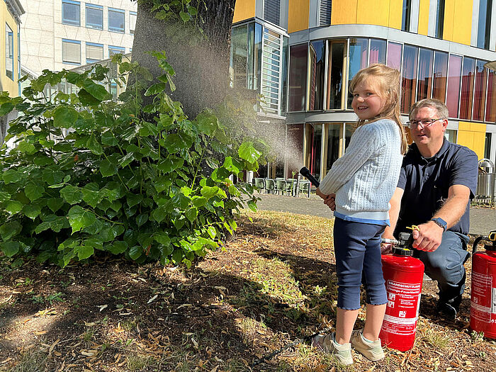 Ein Mädchen und ein Feurwehrmann mit einem Feueröscher vor einem Baum.