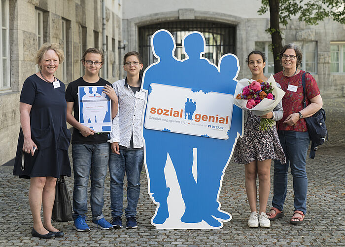 2 Schüler und eine Schülerin stehen, eingerahmt von zwei Lehrerinnen, auf einem Hof. Zwischen den Schülern steht ein Pappaufsteller mit der Aufschrift "Sozial Genial". Die Schülerin hält einen Blumenstrauß in der Hand und ein Schüler hält eine Auszeichnung hoch.