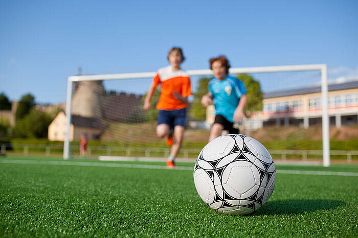 Zu sehen ist ein Ausschnitt eines Fußbaldfeldes. Vorne im Gras liegt der Fußball. Der Hintergrund ist unscharf. Zwei Kinder laufen auf den Ball zu. Das linke Kuind trägt ein orangefarbenes Oberteil. Das rechte Kind, dass auch etwas kleiner ist, trägt ein blaues Oberteil. Beide Kinder sind gleichauf. Hinter ihnen ist ein Fusballtor zu erkennen.