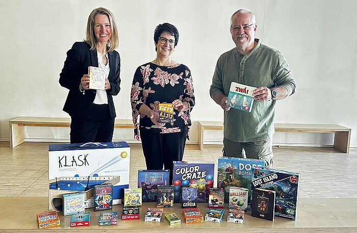 zwei Frauen und ein Mann stehen hinter einem Tisch voller Gesellschaftsspielen.