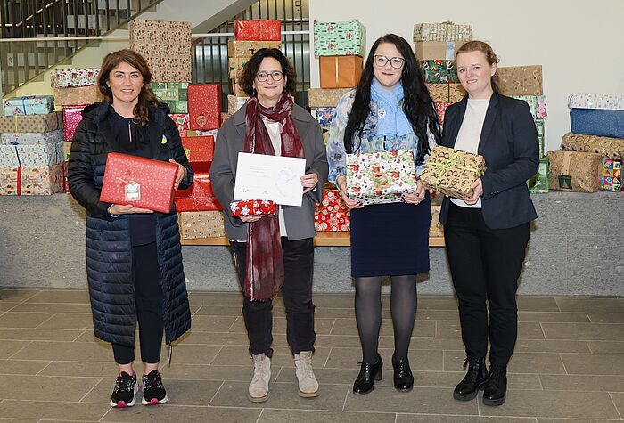 Auf dem Bild stehen Mitarbeiterinnen der StädteRegion und des Vereins Blau-Gelbes Kreuz e.V. vor einem Berg Geschenke und halten repräsentativ jede eins in Händen