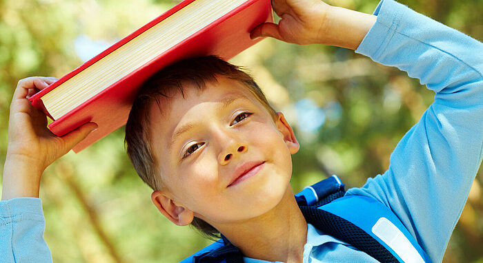 Junge mit Schulranzen trägt Buch auf dem Kopf