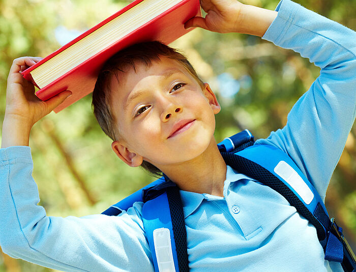 Junge mit Schulranzen trägt Buch auf dem Kopf