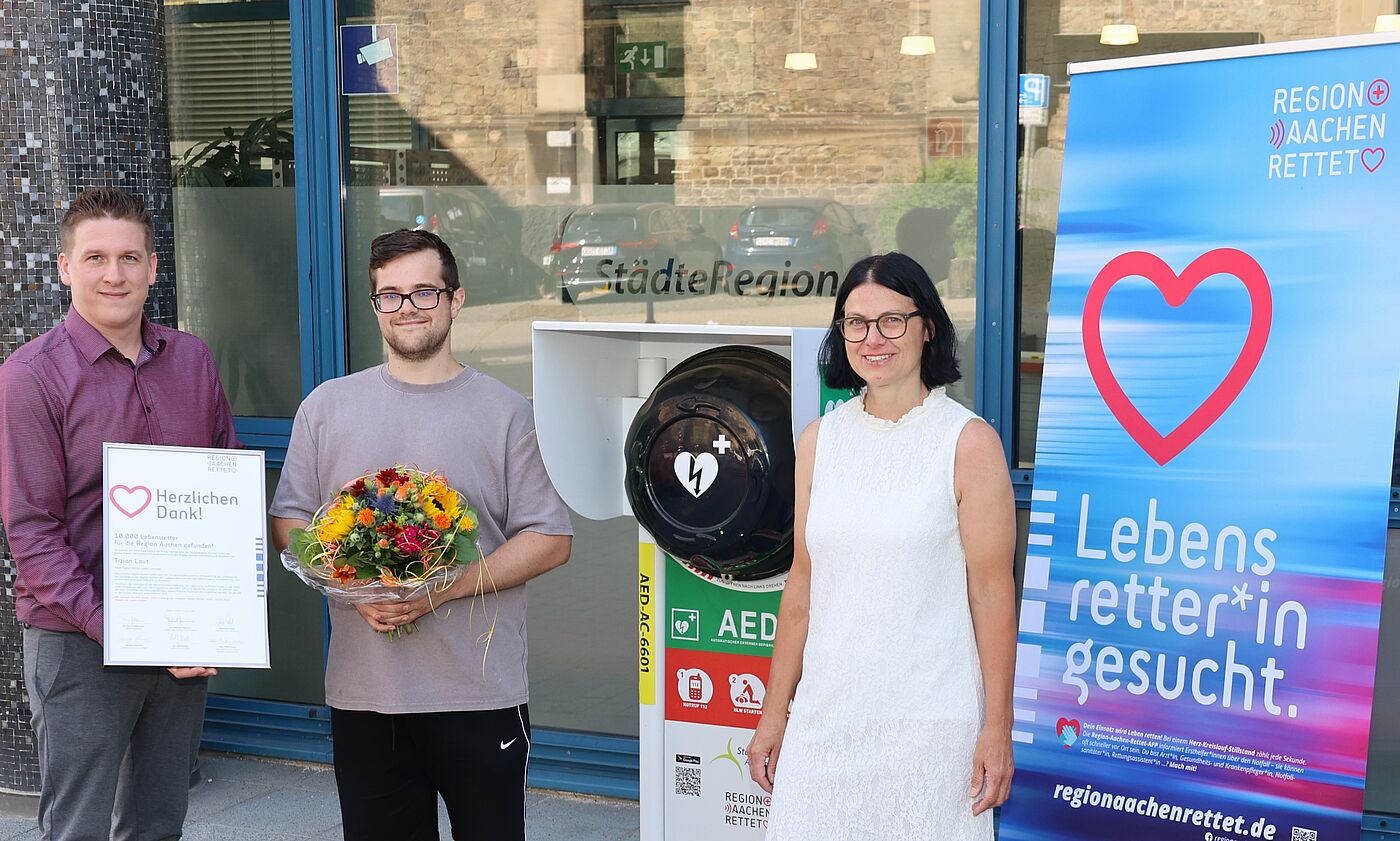 Drei Leute mit Urkunde und Blumenstraß vor einem Defibrillator.