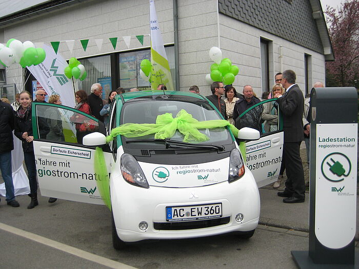 Weißer Smart mit Werbung von EWV Wasserversorgung. Rechts steht eine Ladestation und hinter dem Auto befinden sich mehrere Pwersonen mit grünen und weißen Luftballons inder Hand.