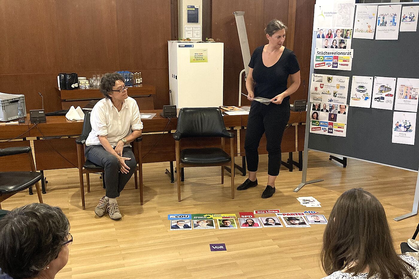 Mehrere Leute sitzen im Kreis und hören einer Frau zu, die etwas zu Bildern auf dem Boden erklärt.