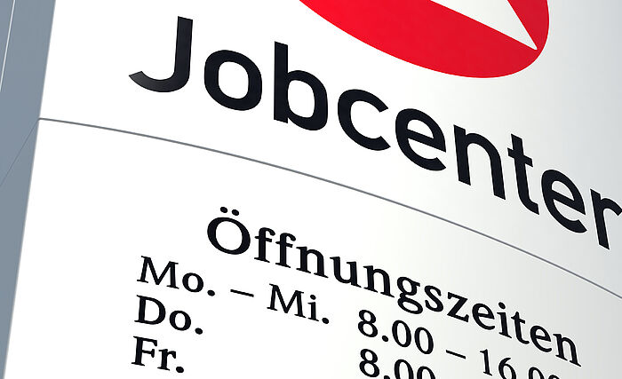 Zu sehen ist ein Schild des Jobcenters. Das Schild beinhaltet neben der Bezeichung auch noch Teile der Öffnungszeit.